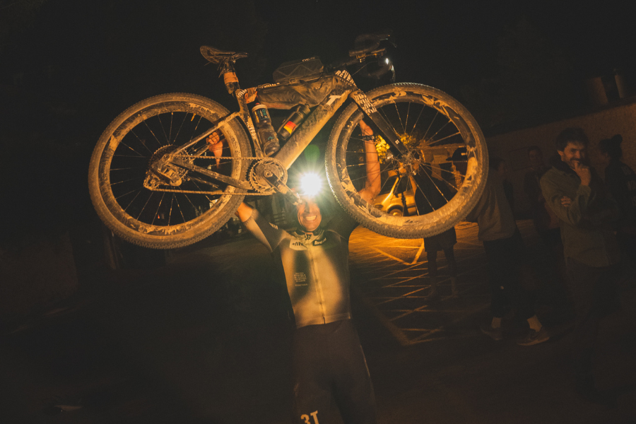 Ekstremni biciklist podiže bicikl iznad glave nakon noćne utrke, ilustracija uspješne energetske strategije i metaboličke izdržljivosti.